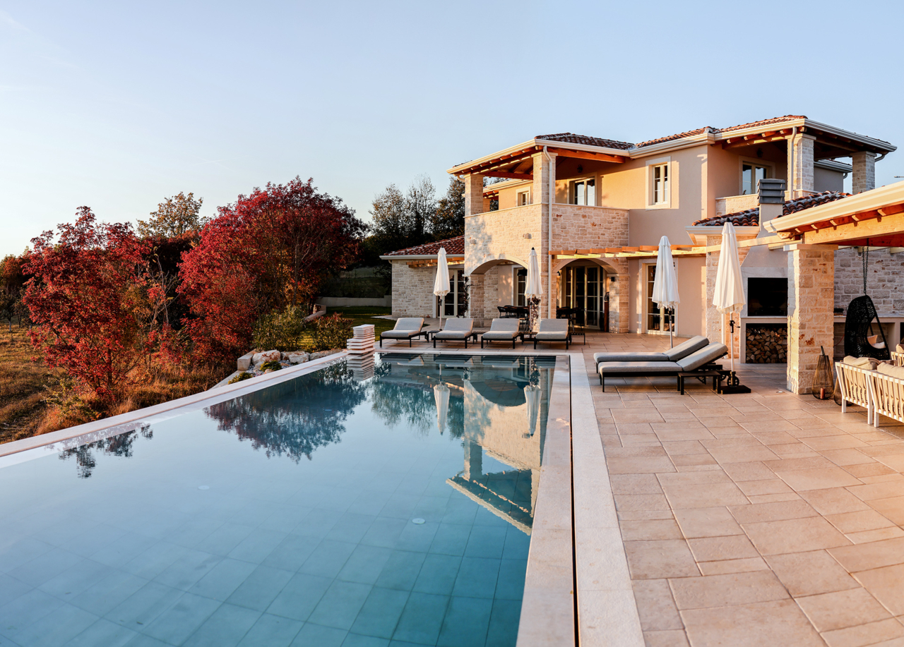 Eine luxuriöse Villa mit einer Terrasse mit Blick auf das umliegende Grün und die Bäume sowie einem Swimmingpool, Sonnenliegen und Sonnenschirm.