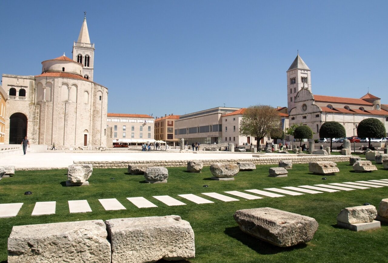Die Überreste des römischen Hauptplatzes mit dem alten Stadtzentrum im Hintergrund und einer Rasenfläche mit Steinresten im Vordergrund.