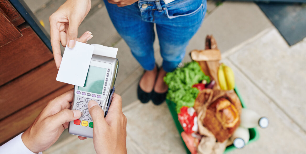 Eine Frau bezahlt mit einer Kreditkarte für Lebensmittel, die in einem Korb auf dem Boden liegen