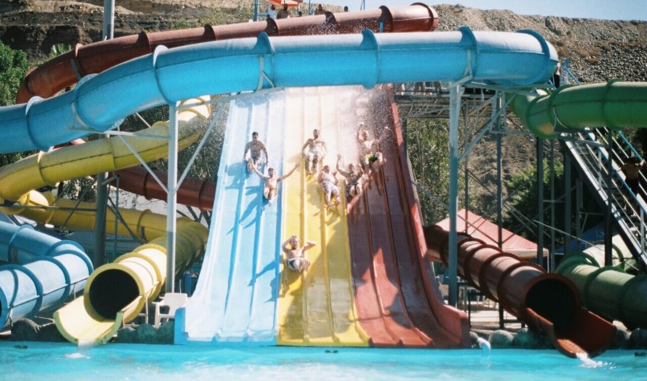 A wide water slide at a water park pool with seven young people sliding down with their hands up in excitement 