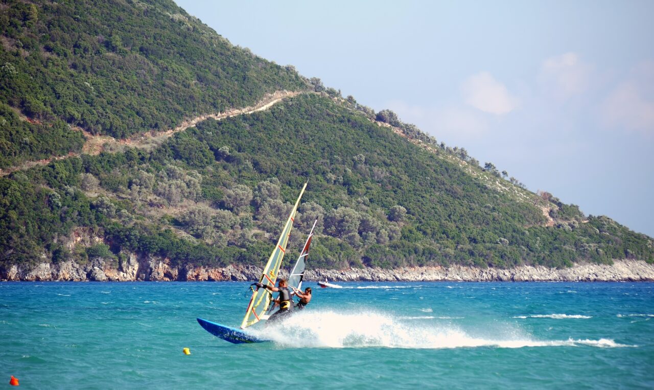 Zwei Windsurfer surfen auf dem Meer, im Hintergrund eine felsige Mittelmeerküste und ein bewaldeter Hügel