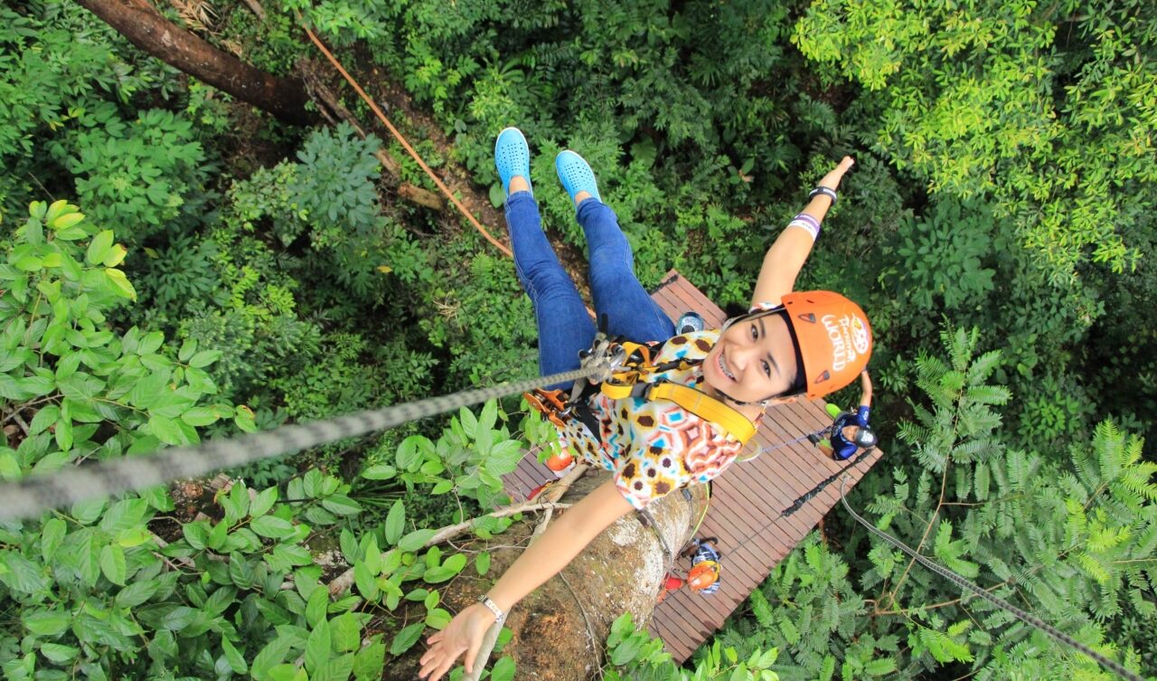 A smiling girl with zip line equipment from a bird's eye view, trees below her